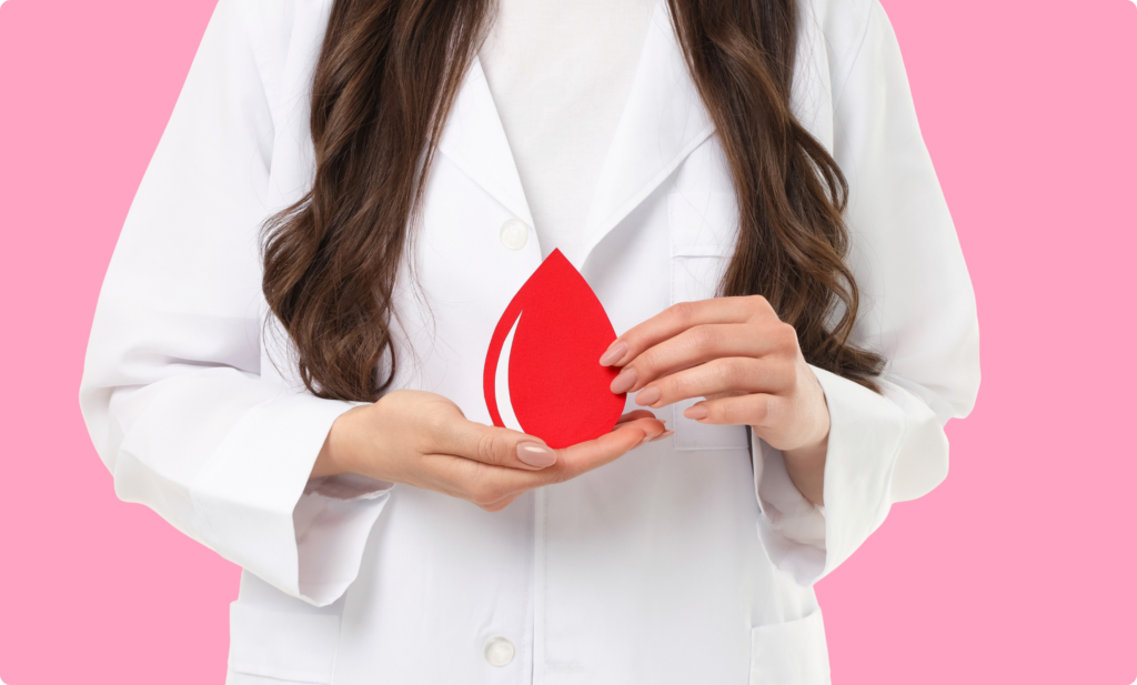 Uma mulher vestindo uma bata branca segura um recorte de papel vermelho em forma de gota de sangue à frente do corpo, contra um fundo rosa sólido. Ela segura a gota com ambas as mãos, posicionada no centro da imagem. O foco está nas mãos e no símbolo da gota de sangue, representando temas de saúde, doação de sangue ou hematologia.